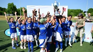 Eine Kinderfußballmannschaft in blauen Trikots des FC Schalke 04 jubelt auf einem Fußballplatz und hält gemeinsam einen großen Pokal in die Höhe. Die Kinder lachen und reißen die Arme in die Luft. Im Hintergrund stehen Erwachsene, die ebenfalls applaudieren und sich mitfreuen. Es ist sonnig und die Stimmung ist ausgelassen und feierlich.
