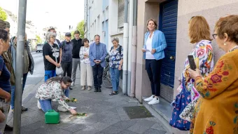 Eine Gruppe von Menschen versammelt sich andächtig auf einem Gehweg vor einem Wohnhaus. Eine Frau kniet nieder und reinigt oder schmückt Stolpersteine mit Blumen, während Christina Rühl-Hamers eine Rede hält.